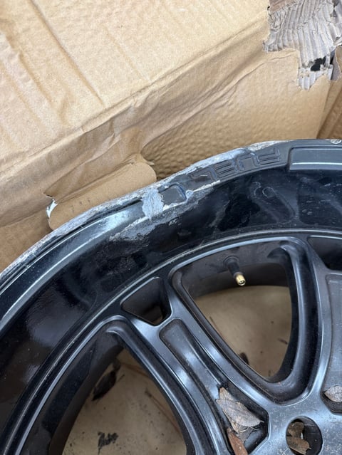 Close-up of a dark wheel rim with visible damage and wear, resting on cardboard packaging material